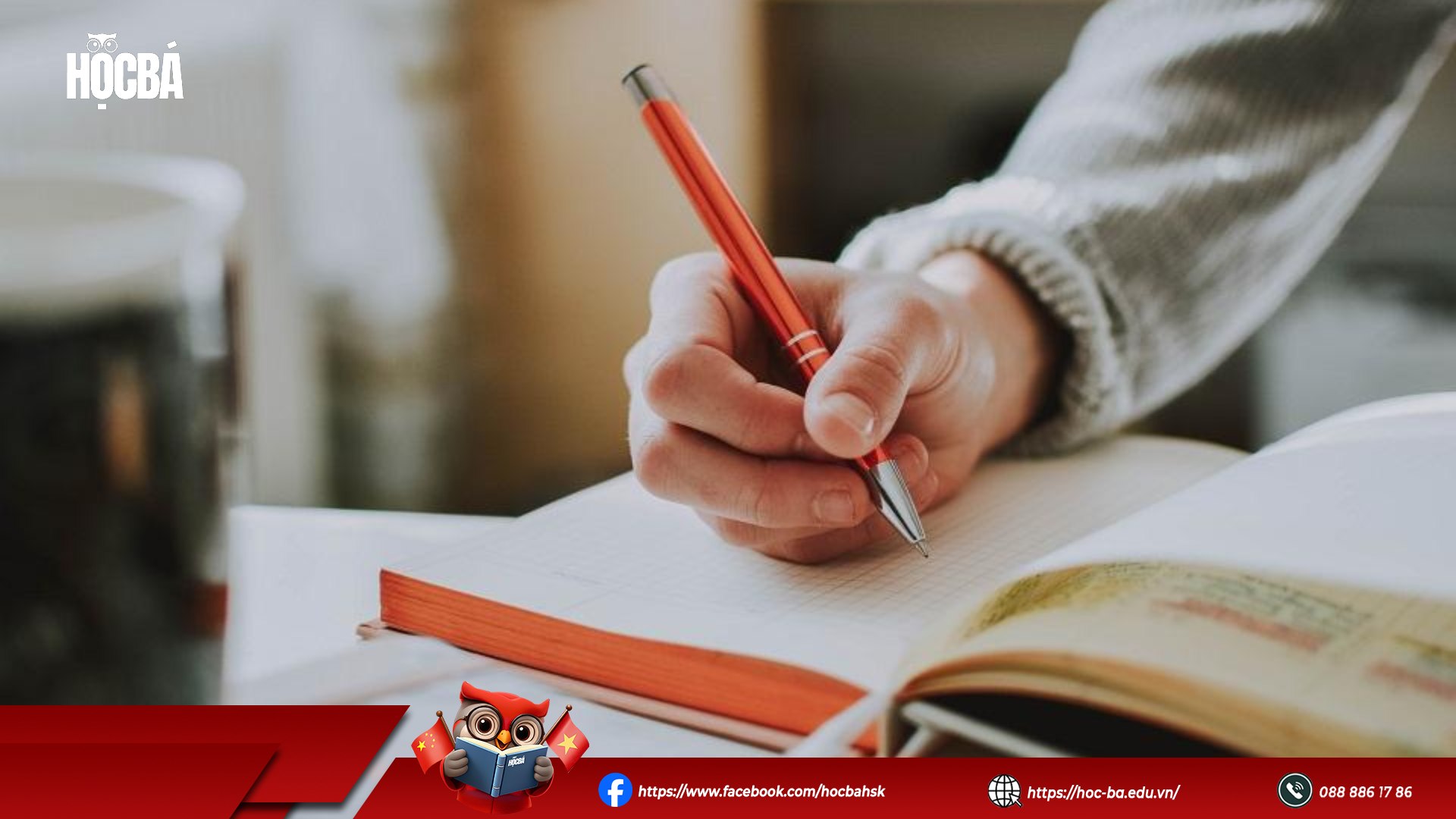 Tay người cầm bút đỏ đang viết vào cuốn vở ô ly bên cạnh một cuốn sách tiếng Trung có in chữ Hán